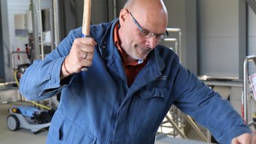 Jürgen Bothner in Blaumann vor Werkplatz schwingt einen Hammer in die Luft, guckt konzentriert.