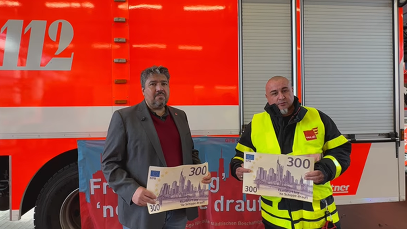 2 Männer vor Feuerwehrauto mit riesigen 300-Euro-Scheinen vor der Brust.